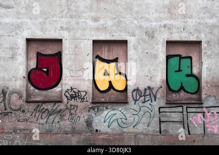 Farbenfrohe Graffiti-Buchstaben, die in alten Fensteraussparungen an einer verwitterten Betonwand gemalt sind, mit zusätzlichen Tags darunter. Stockfoto