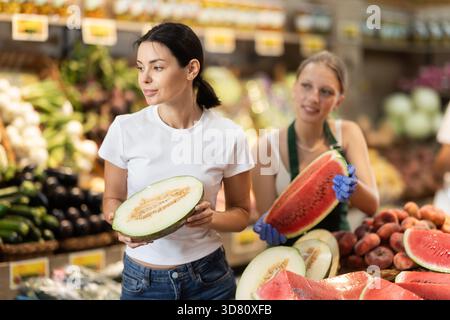 Mädchen wählt reife Melonen im Supermarkt. Weibliche Verkäuferin bietet Wassermelone an Stockfoto