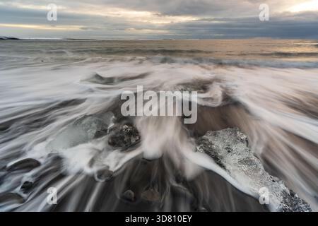 Blick auf Gletschereisfragmente, die auf dem schwarzen Sandstrand verstreut sind, im Kontrast zu den schaumigen weißen Wellen unter einem stimmungsvollen Himmel, Diamond Beach, Sveitarfelag Stockfoto