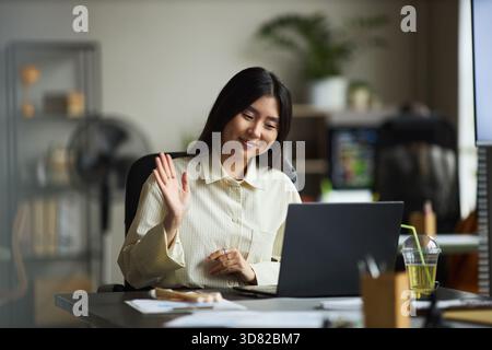 Junge Erwachsene asiatische Frau, die am Schreibtisch sitzt und die Hand winkt, während Videokonferenzen auf einem Laptop in einem modernen Büro stattfinden, lächelt und sich an der Fernkommunikation mit Kollegen beteiligt Stockfoto