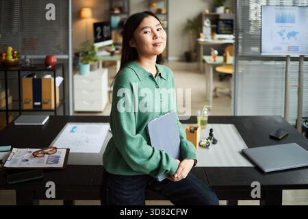 Porträt einer jungen erwachsenen asiatischen Frau, die in einem modernen Büro steht und selbstbewusst Kamera, Geschäftsdokumente und digitale Geräte im Hintergrund auf dem Schreibtisch ansieht Stockfoto