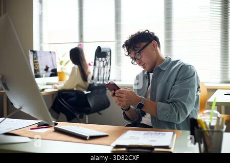 Junger asiatischer Mann, der mit dem Smartphone am Schreibtisch sitzt, während junge asiatische Frau am Computer in modernen Büroarbeitsplätzen arbeitet, beide konzentrierten sich auf Geschäftsaufgaben und Technologie Stockfoto