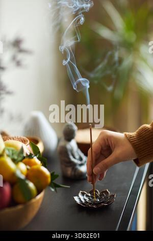 Asiatische junge Erwachsene Frau, die Weihrauchstäbchen in Lotusform mit aufsteigendem Rauch, Obstschale und kleine Buddha-Statue auf dem Tisch, Inneneinrichtung mit verschwommenen Pflanzen Stockfoto