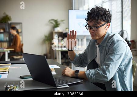 Junger asiatischer Mann mit Brille, der am Schreibtisch sitzt, mit Laptop die Hand hebt, während er an Videokonferenzen im modernen Büro teilnimmt, Geschäftsfrau, die im Hintergrund arbeitet Stockfoto