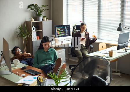 Junge asiatische Geschäftsmänner und Geschäftsfrauen, die in modernen Büros arbeiten, junge asiatische Frau, die am Schreibtisch sitzt und den Computer benutzt, junger asiatischer Mann, der sich mit Kopfhörern entspannt, junge asiatische Frau mit Smartphone Stockfoto