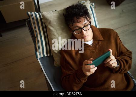 Junger erwachsener asiatischer Mann, der mit dem Smartphone auf der Couch liegt, Brille und lässige Kleidung trägt, sich in moderner Büroumgebung entspannt, sich auf den Bildschirm des Geräts konzentriert und das Telefon mit beiden Händen hält Stockfoto