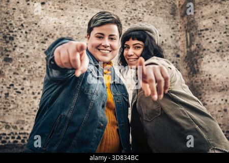 Zwei junge Frauen zeigen mit fröhlichem Ausdruck auf die Kamera und teilen einen Moment der Verbindung und Positivität im Freien. Stockfoto