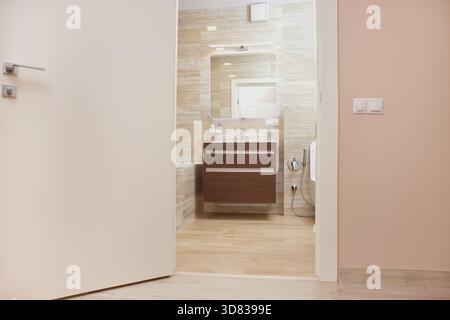 Modernes Bad mit Kosmetikbereich, Waschbecken, Spiegel und gefliesten Wänden, die durch eine offene Tür gesehen werden Stockfoto