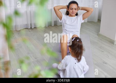 Mutter macht Crunches auf einer Yogamatte, während ihre Tochter in der Nähe beobachtet Stockfoto