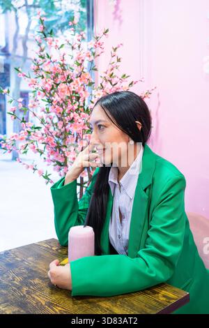 Junge Frau in grünem Blazer, die an einem Café-Tisch mit rosa Hintergrund und Blüten sitzt Stockfoto