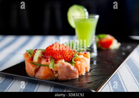 Gourmet-Lachs-Tartar mit leuchtendem rotem Kaviar auf einer schwarzen Platte. Serviert mit frischer Gurke, Dill und einem grünen Shooter-Cocktail. Stockfoto