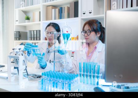 Zwei Wissenschaftlerinnen in weißen Labormänteln arbeiten in einem hellen Labor mit Mikroskopen, Glaswaren und blauen Reagenzgläsern zusammen, die Teamwork repräsentieren Stockfoto