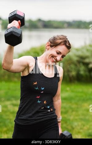 Lächelnde Frau, die Hantel am See in einem grasbewachsenen Park hebt Stockfoto