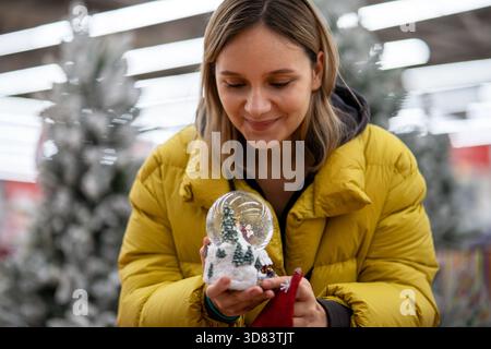 Junge weiße Frau, die eine Schneekugel hält, lächelt beim Einkaufen in einem festlichen Laden mit verschwommenen Weihnachtsbäumen und warmen Lichtern, mit gelbem Puffer Jack Stockfoto