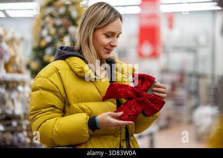 Weiße Frau, die einen gelben Mantel hält, lächelnd, während sie den roten Polka-Dot-Bogen im hellen Weihnachtsgeschäft, weiche Bokeh-Lichter und saisonales d untersucht Stockfoto