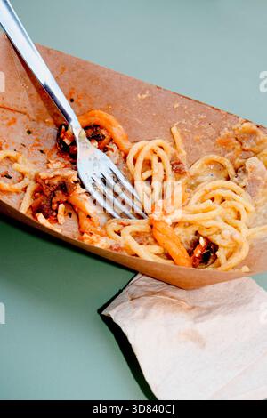 Reste von Casarecce alla norma und Spaghetti alla Carbonara gemischt in einem Einwegkarton mit einer Gabel und einer Serviette auf einem grünen Tisch Stockfoto