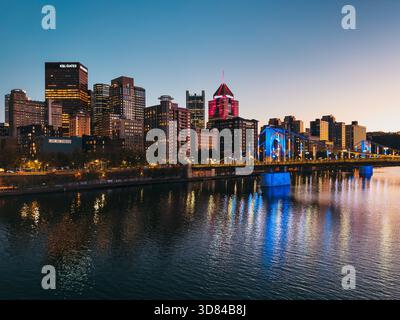 Die Skyline von Pittsburgh in der Abenddämmerung mit beleuchteten Wolkenkratzern und der blau beleuchteten Roberto Clemente Bridge, die sich im Allegheny River spiegelt. Stockfoto