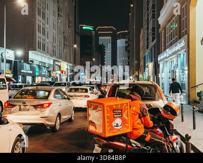 Nächtlicher Verkehr auf der Al Jazeera Street in Doha, Katar, mit einem Talabat-Fahrer inmitten von belebten Autos und beleuchteten Stadttürmen. Stockfoto