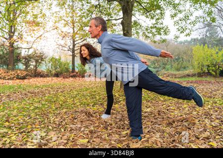 Reifes Paar balanciert auf einem Bein und übt Tai Chi in einem Herbstpark mit gefallenen Blättern Stockfoto