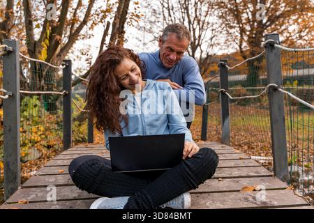 Glückliches Seniorenpaar, das sich einen Laptop auf einer Holzbrücke in einem Herbstpark teilt Stockfoto
