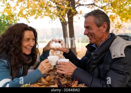 Seniorenpaar, das eine Herzgeste macht, während er Kaffee zwischen Herbstlaub in einem Park genießt Stockfoto