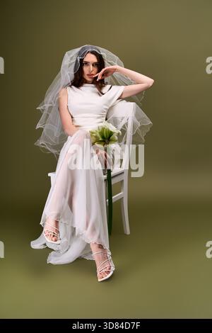 Schöne Braut mit zartem Schleier und Blumenstrauß sitzt elegant und strahlt friedliche Glückseligkeit aus. Stockfoto