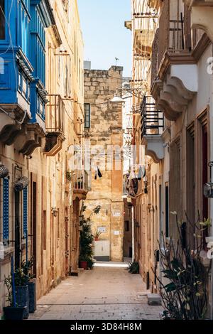 Birgu, Malta? 11. April 2025: Traditionelle schmale maltesische Straße in Birgu mit hellen Kalksteinfassaden, bunten Holzbalkonen, Pflanzen und Wäscherei, die über dem Fußgängerweg hängen Stockfoto