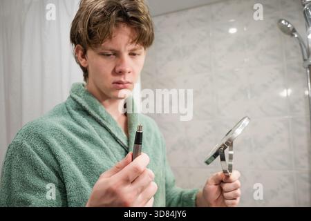 Ein junger kaukasischer Mann, der einen grünen Bademantel trägt, benutzt einen elektrischen Trimmer, um seine Gesichtshaare zu pflegen. Rasierroutine Am Morgen. Nahaufnahme. Stockfoto