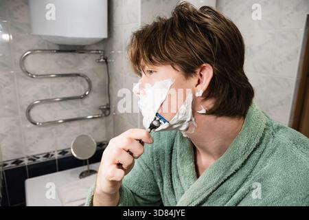 Junger Mann in grünem Gewand trägt Rasiercreme auf und rasiert sich mit Rasierer im Badezimmer. Rasierroutine Am Morgen. Nahaufnahme. Stockfoto