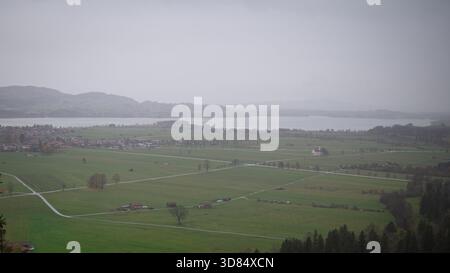 Ein regnerisches ländliches Tal entfaltet sich zu einem See, der von Nebel und niedrigen Wolken weich wird. Die Felder und gewundenen Straßen wirken ruhig unter grauem Himmel. Stockfoto