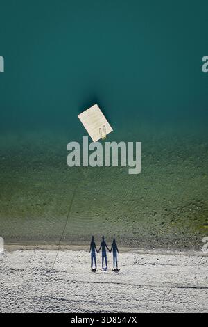 Aus der Vogelperspektive von drei Figuren, die auf einem leichten Sandstrand stehen, der durch eine dünne Linie mit einer schwimmenden Plattform auf dem türkisfarbenen Wasser verbunden ist, Fjord, Norwegen. Stockfoto