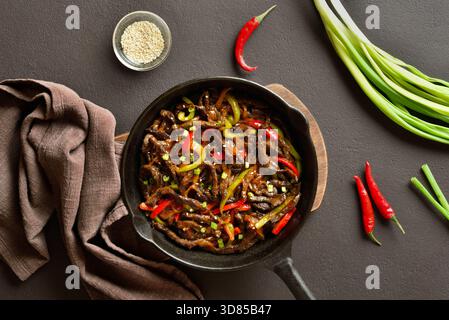 Frittiertes, dünn geschnittenes Rindersteak mit Paprika in gusseiserner Pfanne auf dunkelbraunem Hintergrund. Draufsicht, flach Stockfoto