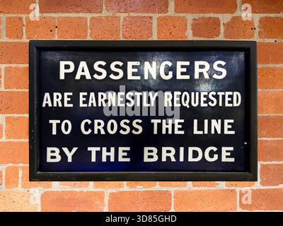 Vintage-Eisenbahnschild, das die Passagiere anweist, die Strecke über die Brücke zu überqueren, die an einer roten Ziegelmauer befestigt ist Stockfoto