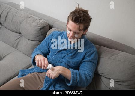Junger kaukasischer Mann im blauen Bademantel, der Hautpflegecreme mit Augenflecken aufträgt, während er während der Selbstpflege zu Hause auf dem Sofa sitzt. Herren Home Self Stockfoto