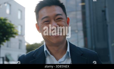 Porträt glücklicher lächelnder asiatischer Geschäftsmann in der Stadt koreanischer Geschäftsmann, der in die Kamera schaut Lächeln zahnig erfreut sich sorgloser chinesischer japanischer Mann mittleren Alters Stockfoto