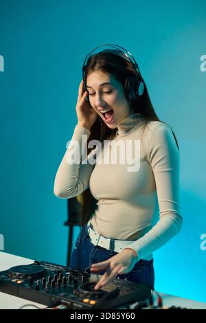 Weibliche DJ-Mischpulte auf Plattenspielern, farbenfrohe Lichtstudio, fokussierter Ausdruck, Hände einstellender Mixer und Laptop-Bedienelemente, Kopfhörer tragen, modern Stockfoto