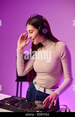 Weibliche DJ-Mischpulte auf Plattenspielern, farbenfrohe Lichtstudio, fokussierter Ausdruck, Hände einstellender Mixer und Laptop-Bedienelemente, Kopfhörer tragen, modern Stockfoto