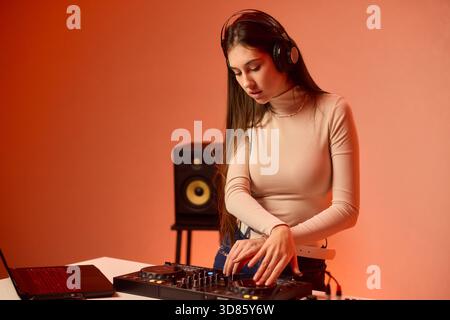 Weibliche DJ-Mischpulte auf Plattenspielern, farbenfrohe Lichtstudio, fokussierter Ausdruck, Hände einstellender Mixer und Laptop-Bedienelemente, Kopfhörer tragen, modern Stockfoto
