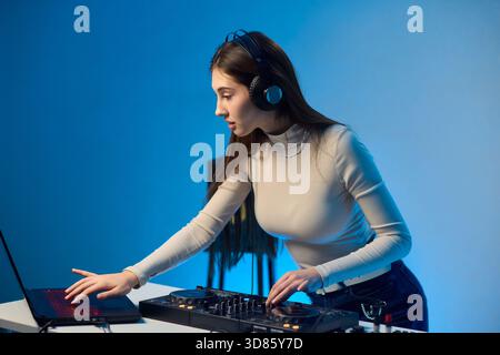 Weibliche DJ-Mischpulte auf Plattenspielern, farbenfrohe Lichtstudio, fokussierter Ausdruck, Hände einstellender Mixer und Laptop-Bedienelemente, Kopfhörer tragen, modern Stockfoto