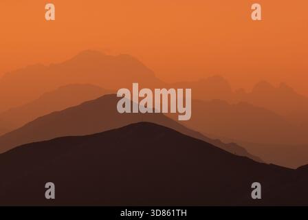 Blick auf die geschichteten Bergsilhouetten, die in den trüben orangefarbenen Himmel fallen und eine ruhige und ätherische Landschaft schaffen, Jamnik, Region Zilina, Slowakei. Stockfoto