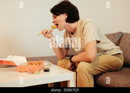 Eine Frau sitzt auf einem Sofa und isst gebratenes Sushi mit Essstäbchen aus einer Lieferbox. Die Szene hebt das zwanglose Essen, den Komfort und das moderne Zuhause hervor Stockfoto