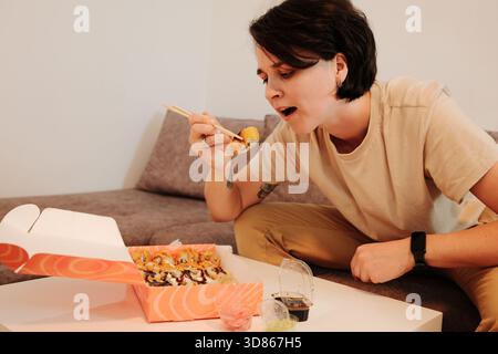 Eine Frau sitzt auf einem Sofa und isst gebratenes Sushi mit Essstäbchen aus einer Lieferbox. Die Szene hebt das zwanglose Essen, den Komfort und das moderne Zuhause hervor Stockfoto