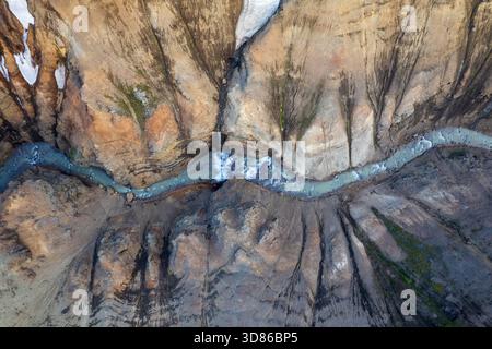 Aus der Vogelperspektive des Gletscherflusses, der durch die zerklüftete, farbenfrohe Landschaft schlängelt, wo Schneestreifen auf die Erdtöne in Kerlingarfjöll, Hrunamann, treffen Stockfoto