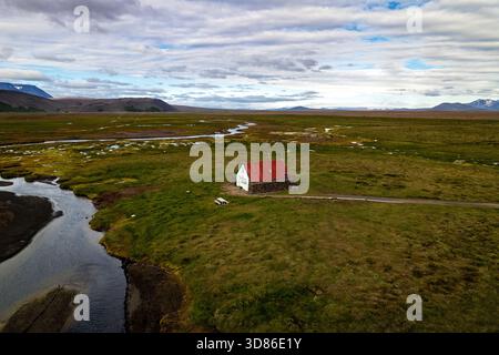 Aus der Vogelperspektive auf ein einsames Haus mit einem roten Dach inmitten einer weiten, grünen Landschaft, die von sich windenden Flüssen unter einem bewölkten Himmel geteilt wird, Hvíta, Island. Stockfoto