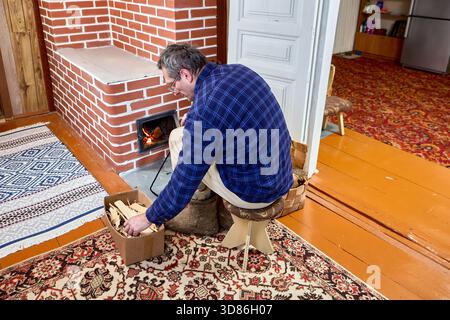 Kamin roter Ziegelholzofen niedriger Hocker vor dem Herd Öffnung Flamme brennender reifer Mann sitzt auf der Suche nach etwas Kiste entzündet. Stockfoto