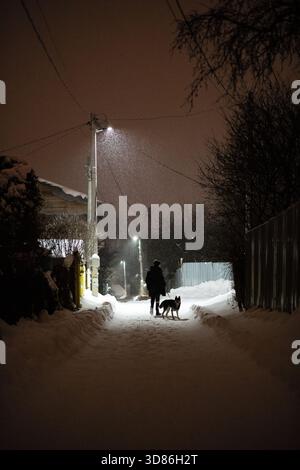 Person, die nachts mit einem Hund auf einer verschneiten Dorfstraße spaziert, beleuchtet von Straßenlaternen. Winterszene in Russland. Stockfoto