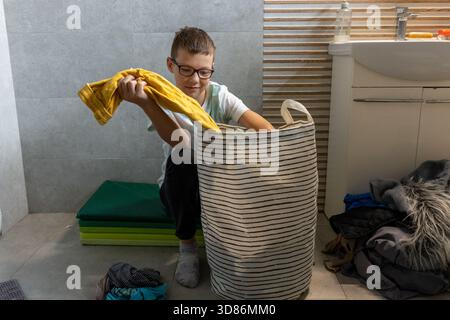 Junge mit Brille, der schmutzige Wäsche in gestreiften Korb im Badezimmer zu Hause sortiert. Das Konzept der Kinder, die bei der Hausarbeit helfen, unterrichten Stockfoto