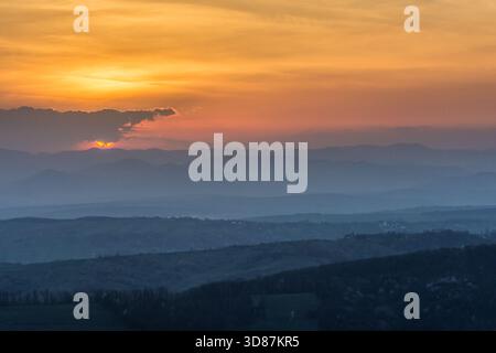 Mehrschichtige Bergkämme, die unter einem dramatischen Sonnenuntergang in die Ferne gleiten. Das warme Leuchten steht im Kontrast zu kühlen Blau- und Violetttönen. Stockfoto