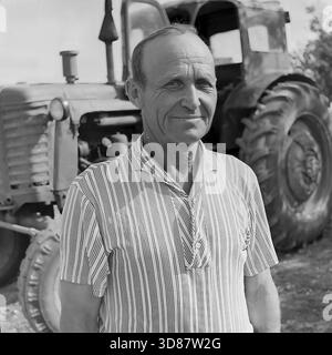 Donbass-Region, Ukraine, UdSSR - August 1979: Porträt eines Traktorfahrers, der vor einem sowjetischen MTZ-50-„Weißrussland“-Traktor im Veselowski-Tonsteinbruch bei Druschkowka steht. Der Mann mittleren Alters trägt ein gestreiftes Kurzarmhemd. Er hat ein gebräuntes, verwittertes Gesicht mit tiefen Falten. Die Landwirtschaftsmaschine hinter ihm verfügt über große Hinterräder und eine offene Kabinentür. Dieses Schwarzweiß-Archivfoto fängt den Charakter der Landarbeiter und die Atmosphäre der friedlichen Donbass-Region ein. Stockfoto