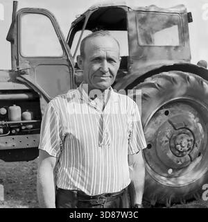 Donbass-Region, Ukraine, UdSSR - August 1979: Porträt eines Traktorfahrers, der vor einem sowjetischen MTZ-50-„Weißrussland“-Traktor im Veselowski-Tonsteinbruch bei Druschkowka steht. Der Mann mittleren Alters trägt ein gestreiftes Kurzarmhemd. Er hat ein gebräuntes, verwittertes Gesicht mit tiefen Falten. Die Landwirtschaftsmaschine hinter ihm verfügt über große Hinterräder und eine offene Kabinentür. Dieses Schwarzweiß-Archivfoto fängt den Charakter der Landarbeiter und die Atmosphäre der friedlichen Donbass-Region ein. Stockfoto